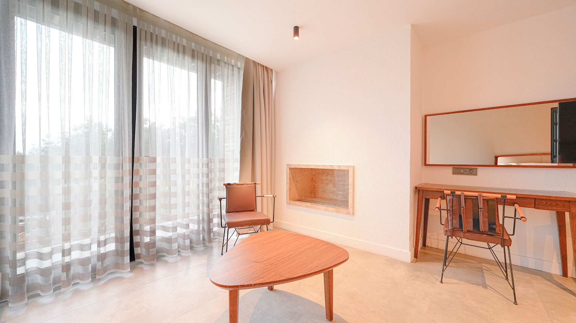 Una habitación de hotel moderna con un ventanal grande, una mesa de madera, una silla, un pequeño banco de madera y un televisor sobre un mueble de madera.