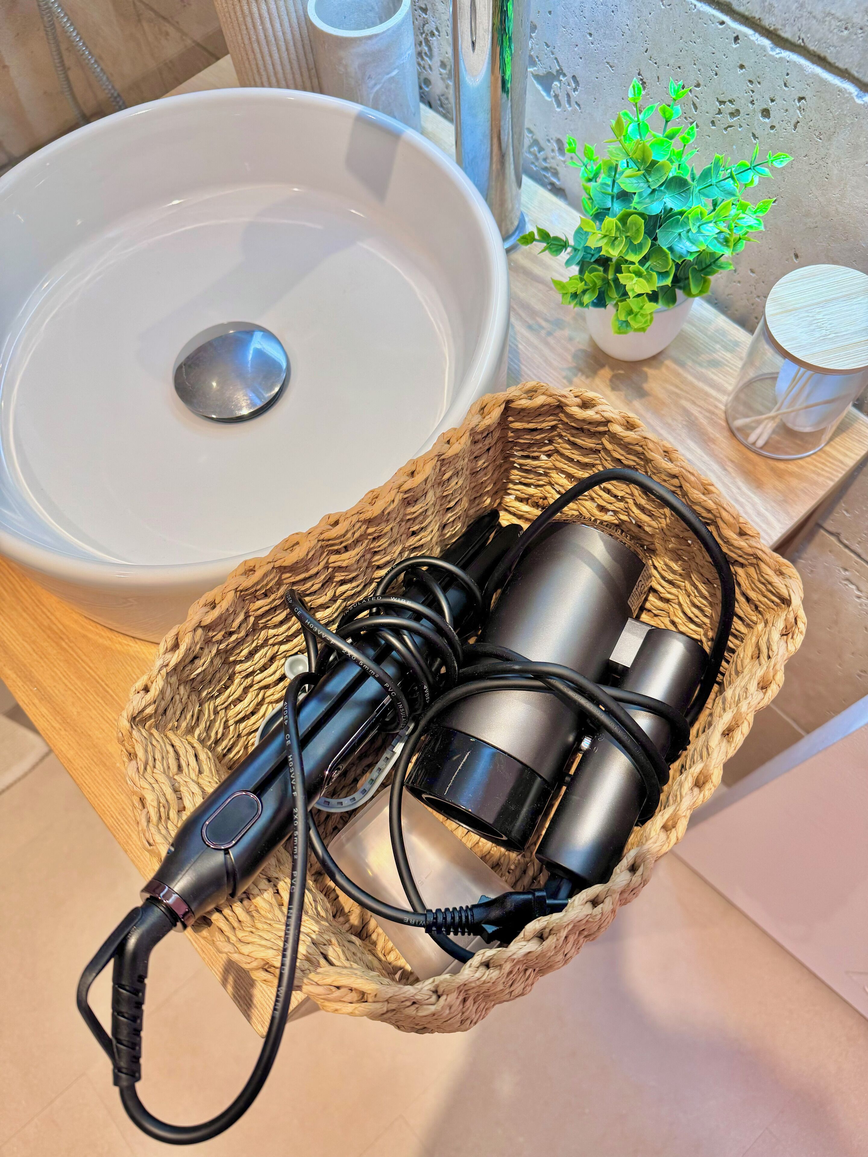 A bathroom with a white sink, a woven basket containing a hairdryer and its cord, a potted plant, and a glass container on a wooden tray.