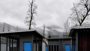Une rangée de cabanes aux portes et fenêtres bleues, entourées d’un mur de pierre et bordées d’un chemin de terre.