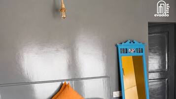 A bed with an orange bedspread and two pillows, a wall-mounted light fixture, and a blue mirror leaning against the wall.