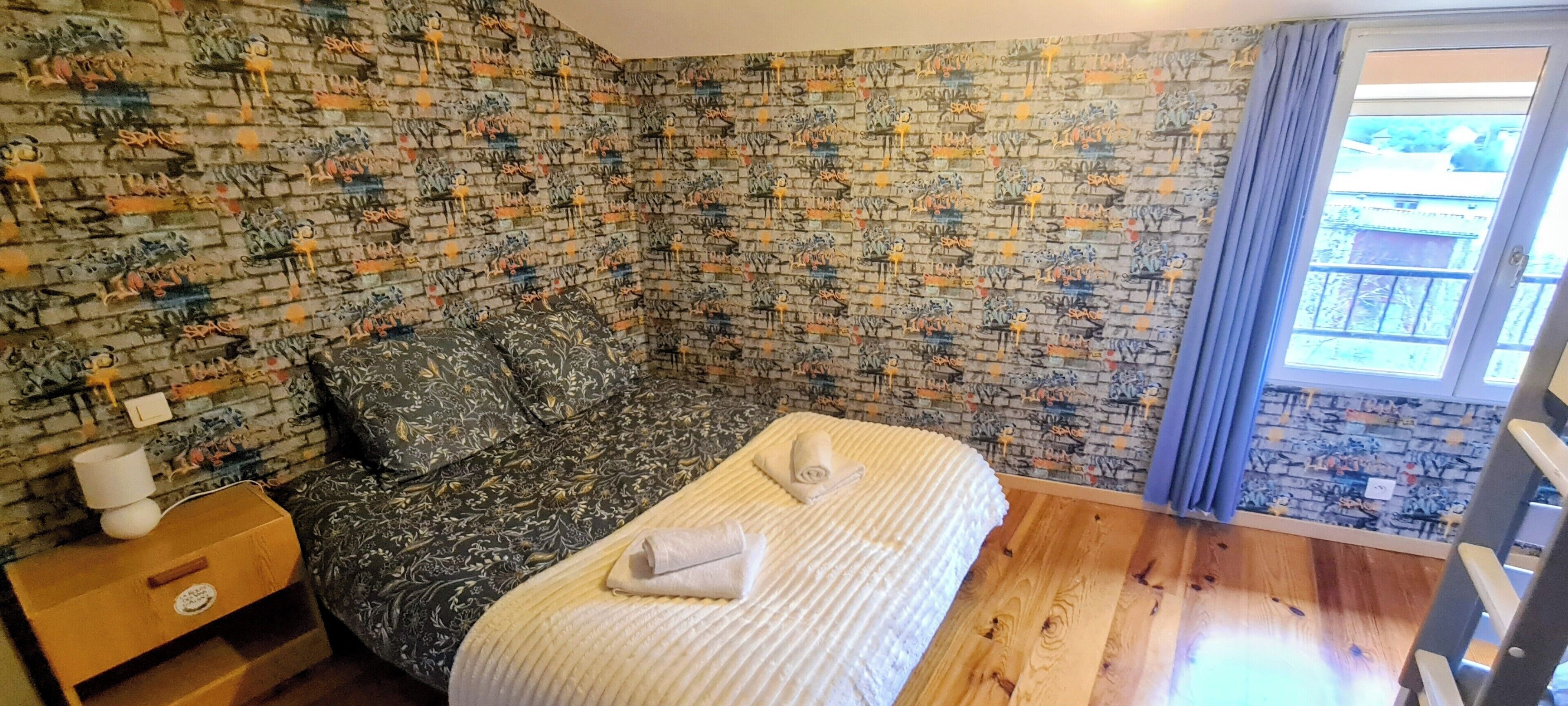 Une chambre avec un mur décoré d’un motif en briques, un lit recouvert d’une couverture blanche, une table de chevet avec une lampe et une fenêtre avec des rideaux.