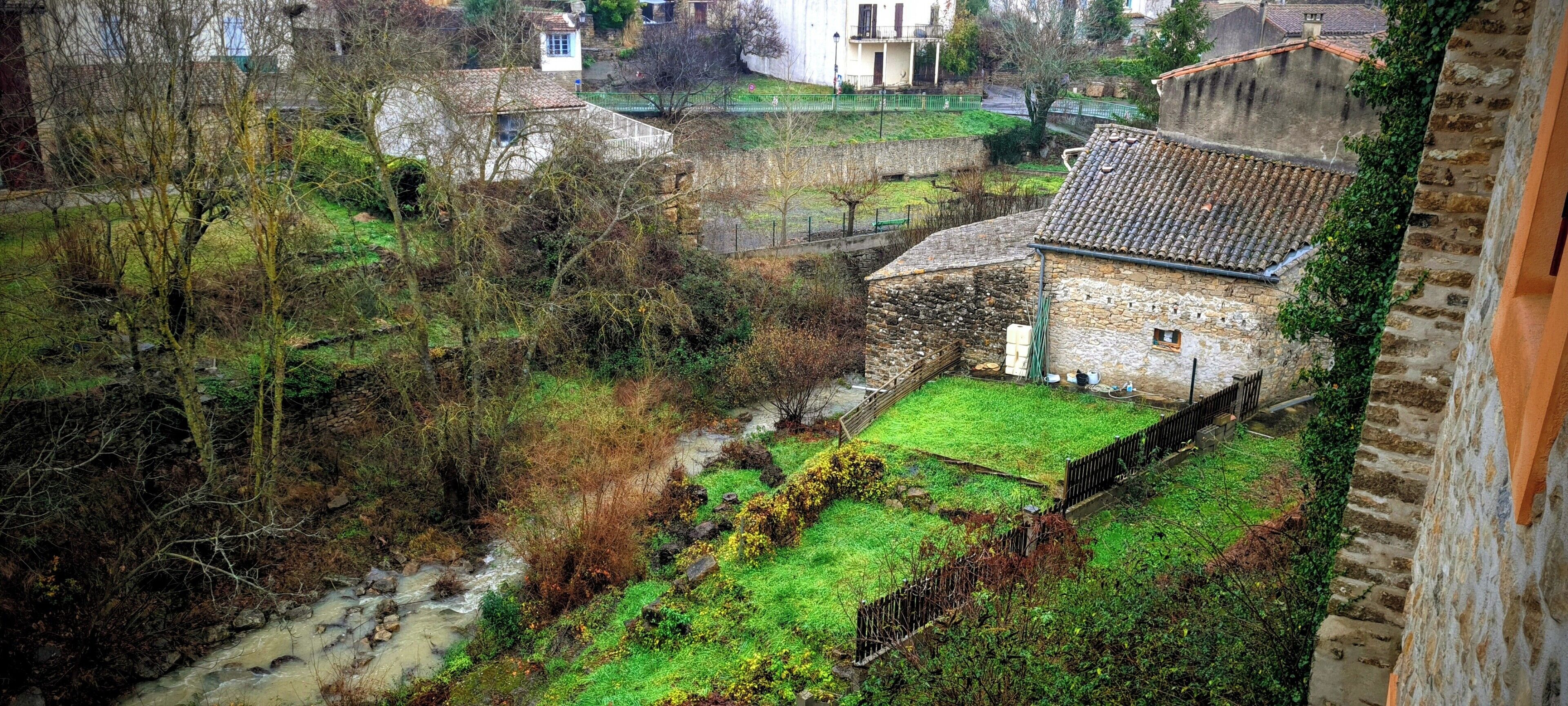 A stone building with a tiled roof, surrounded by greenery and a small stream.
