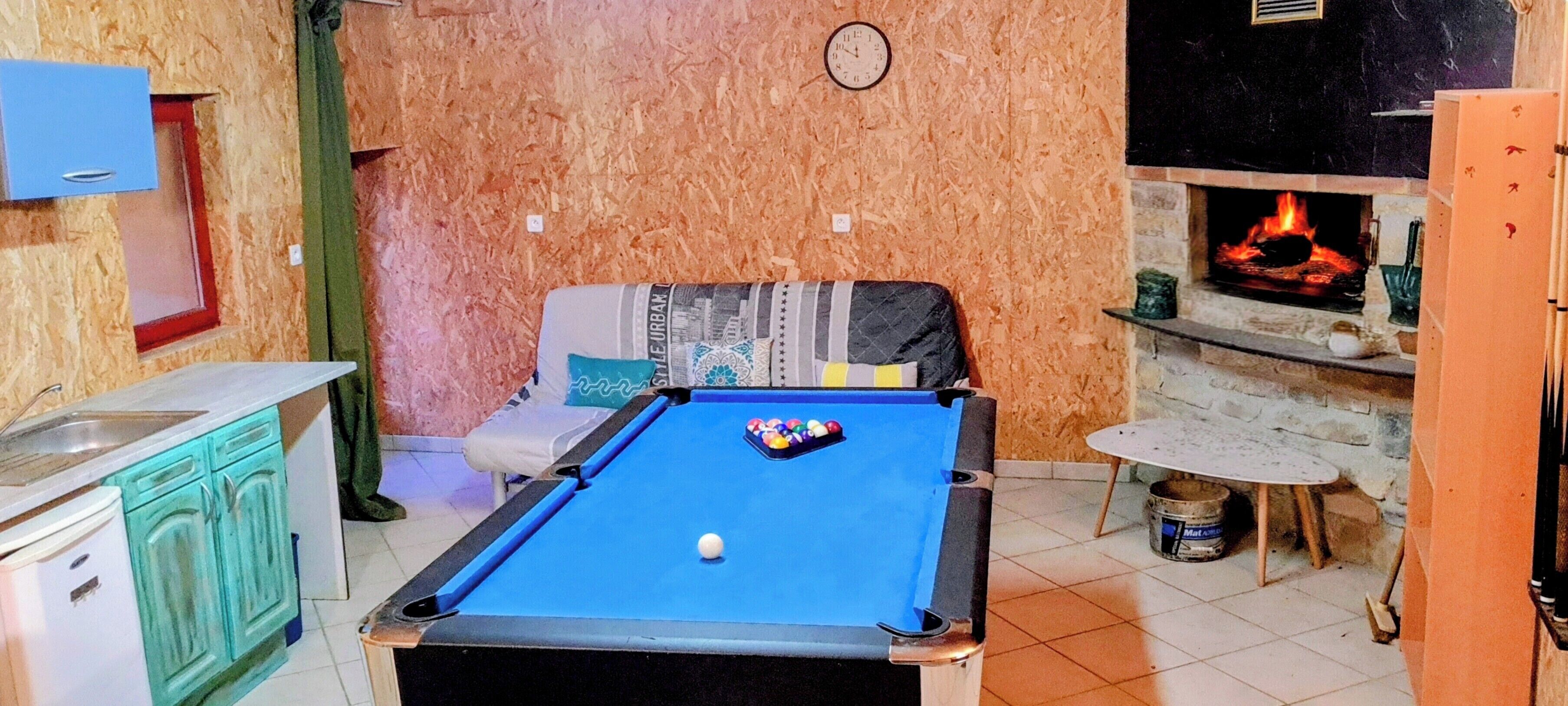 A room with a pool table, a sofa, a small kitchen area, and a fireplace.
