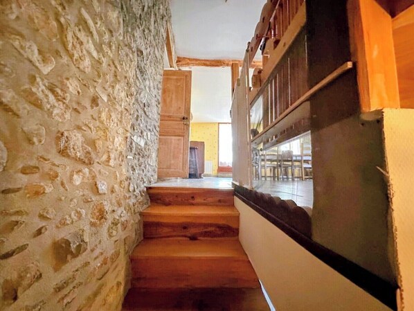 Un escalier étroit aux murs de pierre et aux marches en bois mène à une pièce dotée d’une grande fenêtre et d’un plafond en bois.