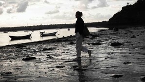 Una persona caminando sobre una playa mojada, con barcos en el agua y un cielo nublado.