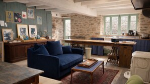 Una sala rústica con un sofá azul, una mesa de centro de madera y un sillón verde. Se ve un área de cocina con una pared de piedra y una ventana.