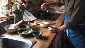Una cocina rústica con una persona cocinando, un muro de piedra, un ventanal con flores y diversos utensilios de cocina.