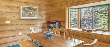 A wooden cabin interior with a dining table, chairs, a bookshelf, and a window with a forest view.
