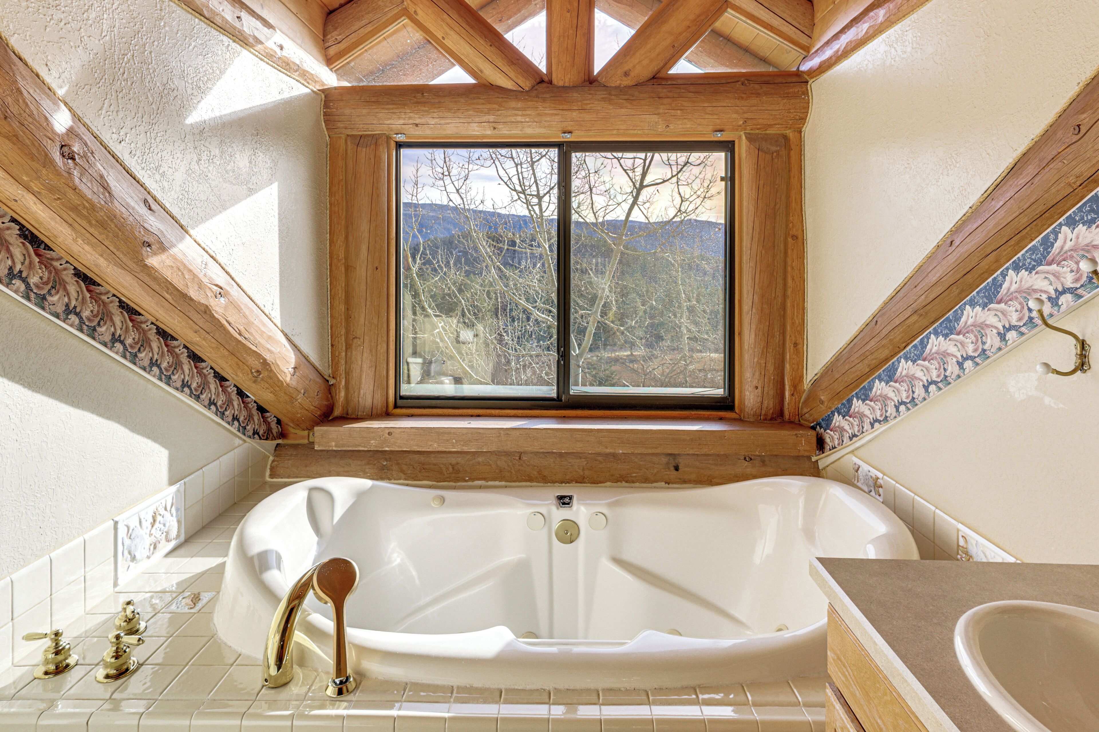 A bathroom with a large bathtub, wooden ceiling beams, and a window offering a view of trees and mountains.