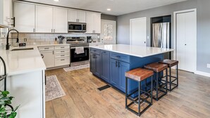 A modern kitchen with white and blue cabinets, a central island with stools, stainless steel appliances, and a wooden floor. - Waterfront Deck, Shell Knob Gem on Table Rock! (Shell Knob)