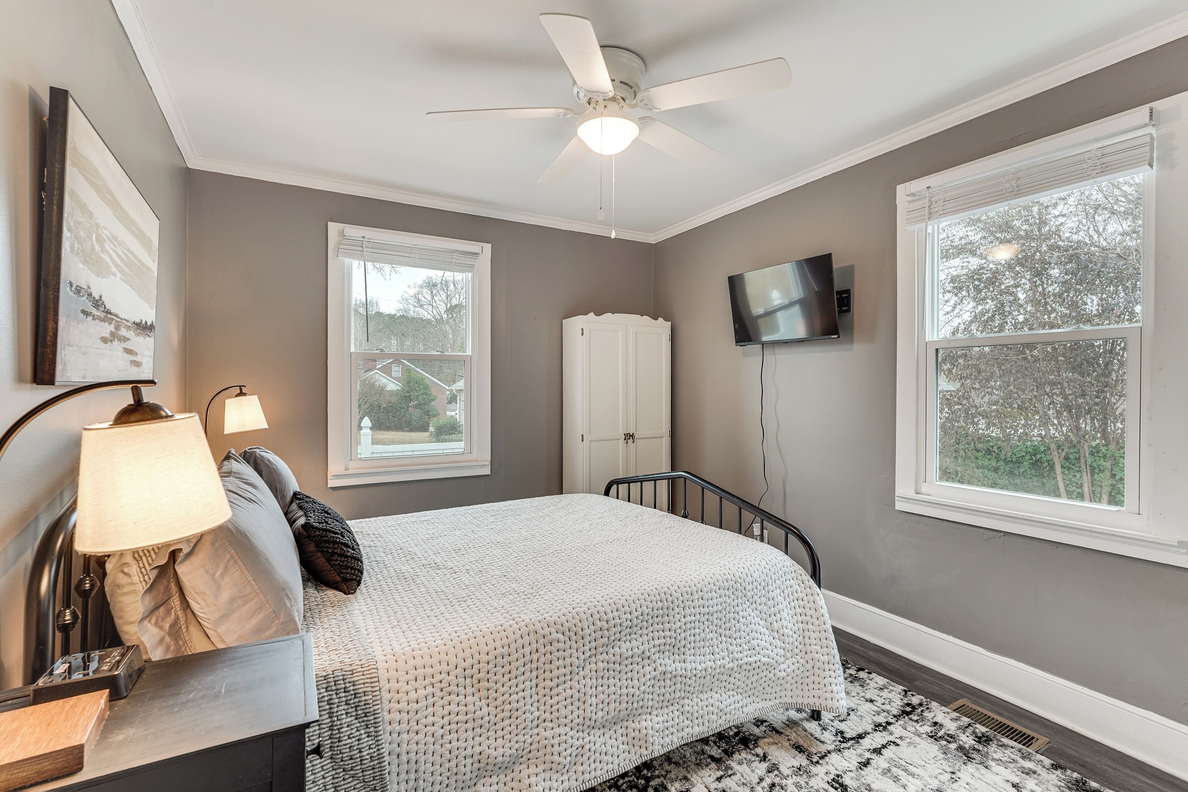 A bedroom with a bed, a ceiling fan, a TV mounted on the wall, and a window.