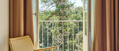 Una habitación con cama, una silla y un balcón con vistas a un árbol alto.
