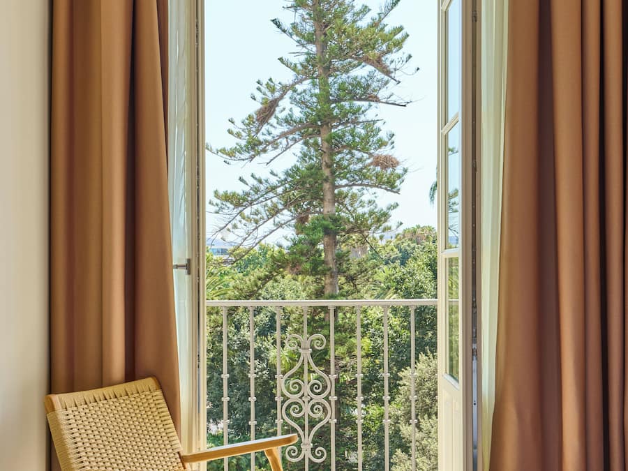 A room with a bed, a chair, and a balcony with a view of a tall tree.