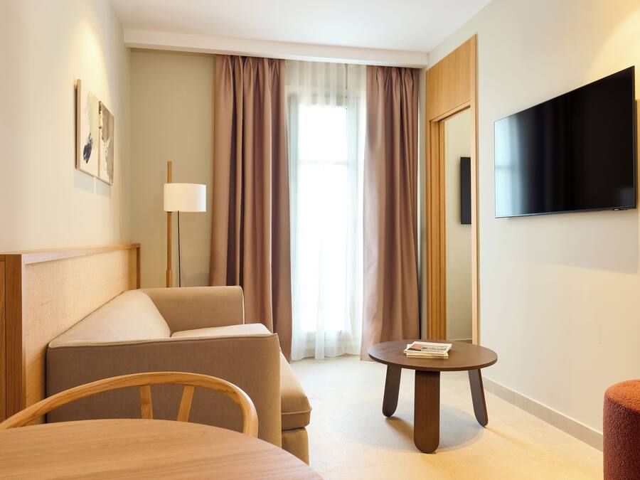Habitación de hotel con cama, sofá, una mesita, televisión y ventana con cortinas.