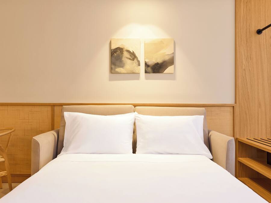 A hotel room with a bed, two pillows, a chair, and two wall-mounted paintings.