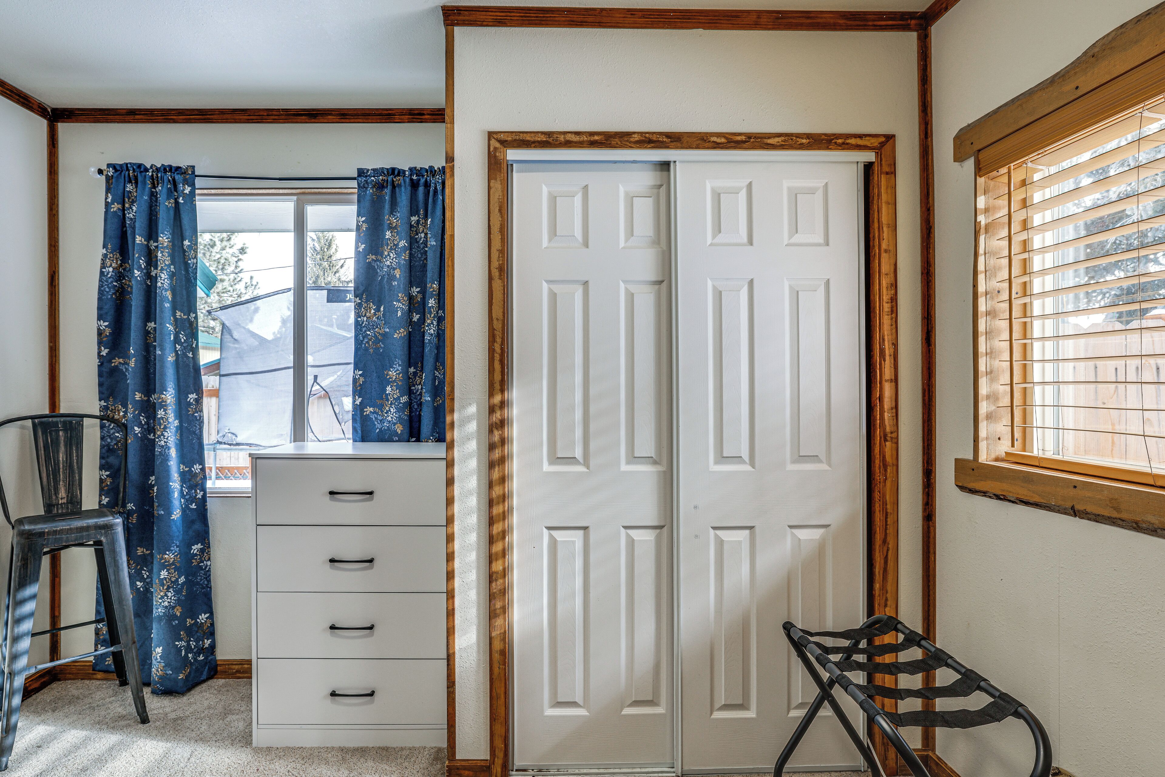 A room with a white door, a desk with drawers, a chair, and a window with blinds.