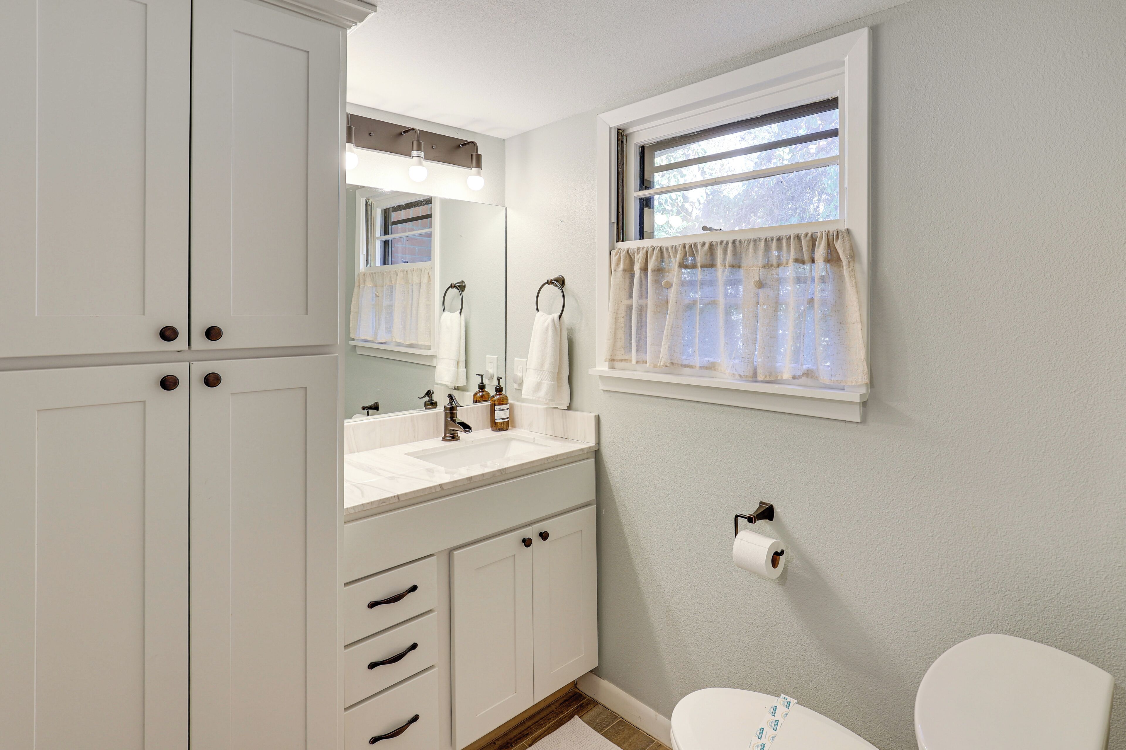 A bathroom with a white sink, mirror, and toilet. There is a window with a curtain, and a towel rack above the sink.