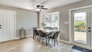 Un coin repas avec une table et des chaises en bois, un ventilateur de plafond et une vue sur l’extérieur, visible à travers une porte.
