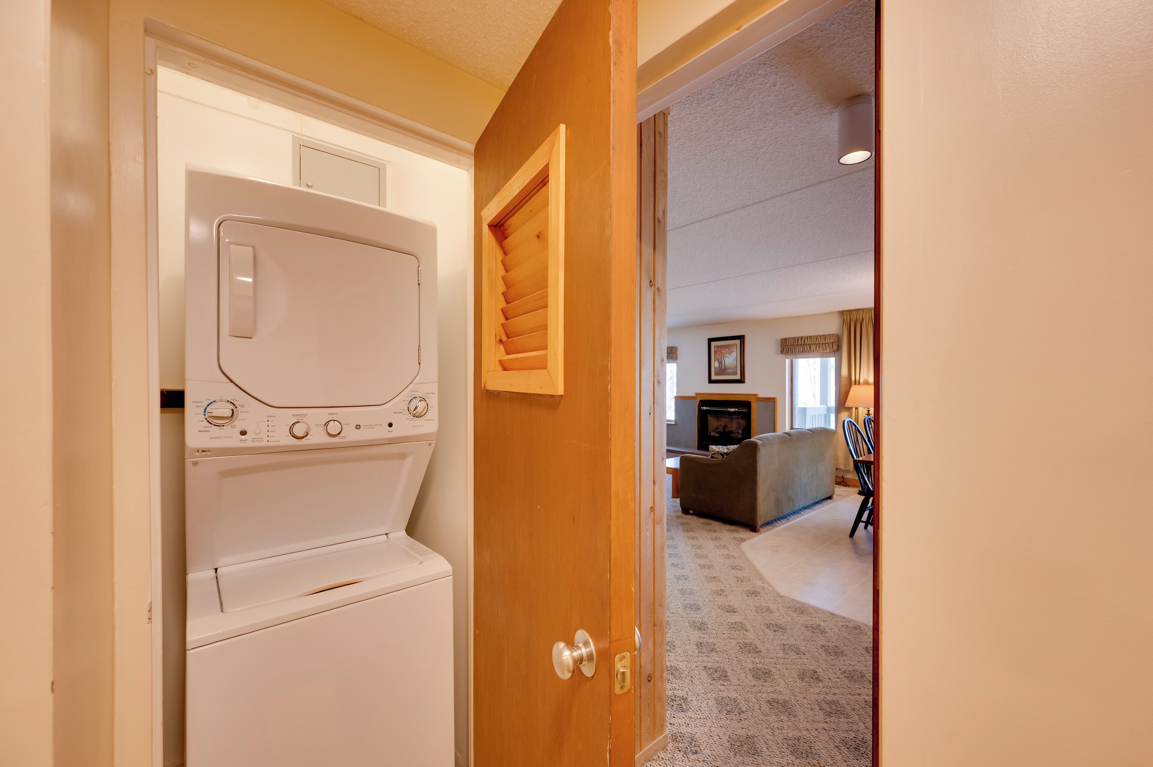 A laundry room with a front-loading washer and dryer, a wooden door, and a living area with a couch, TV, and a small table.