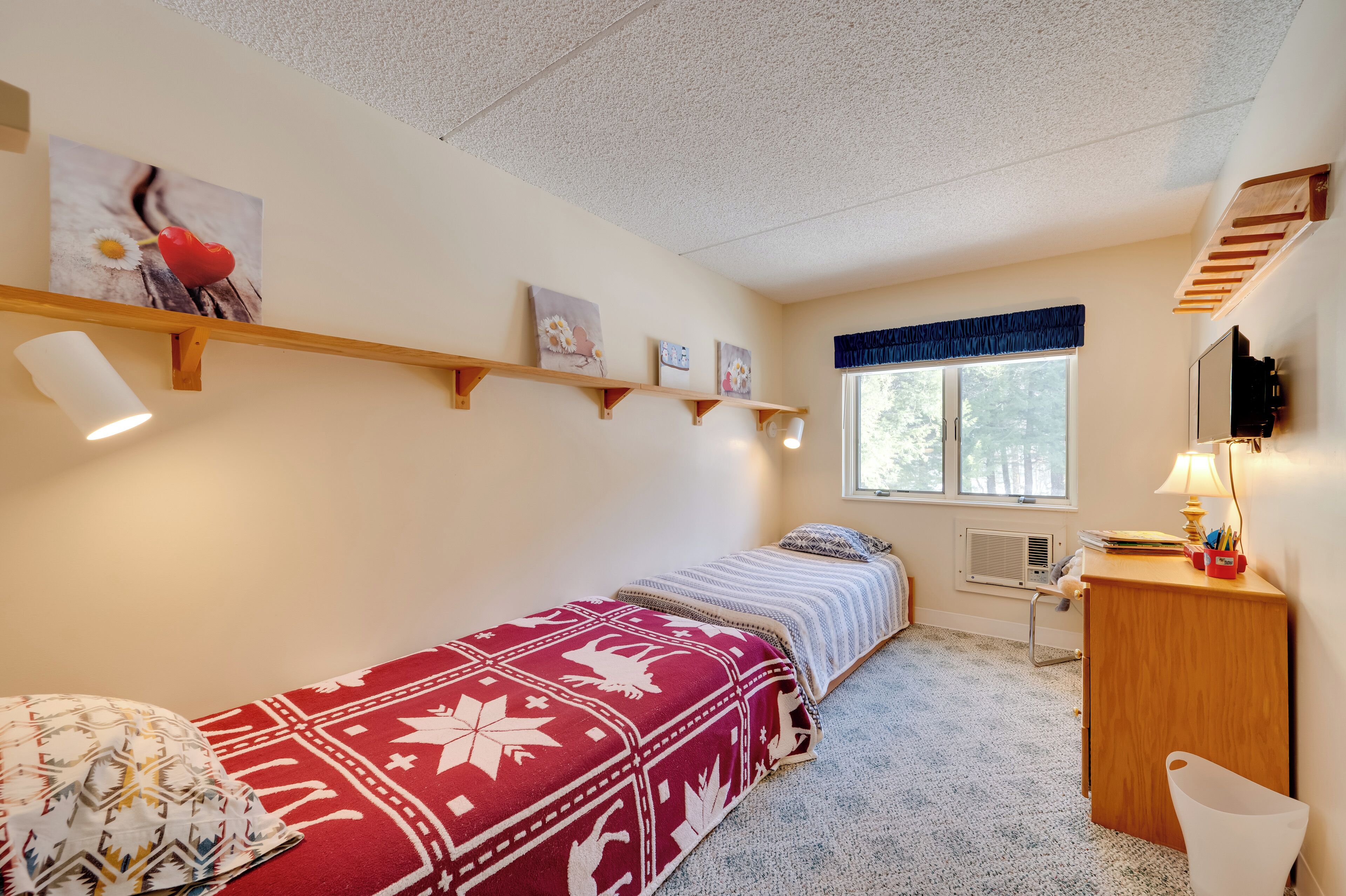 Un dormitorio con cama, escritorio, silla, ventana y estanterías fijas a la pared.