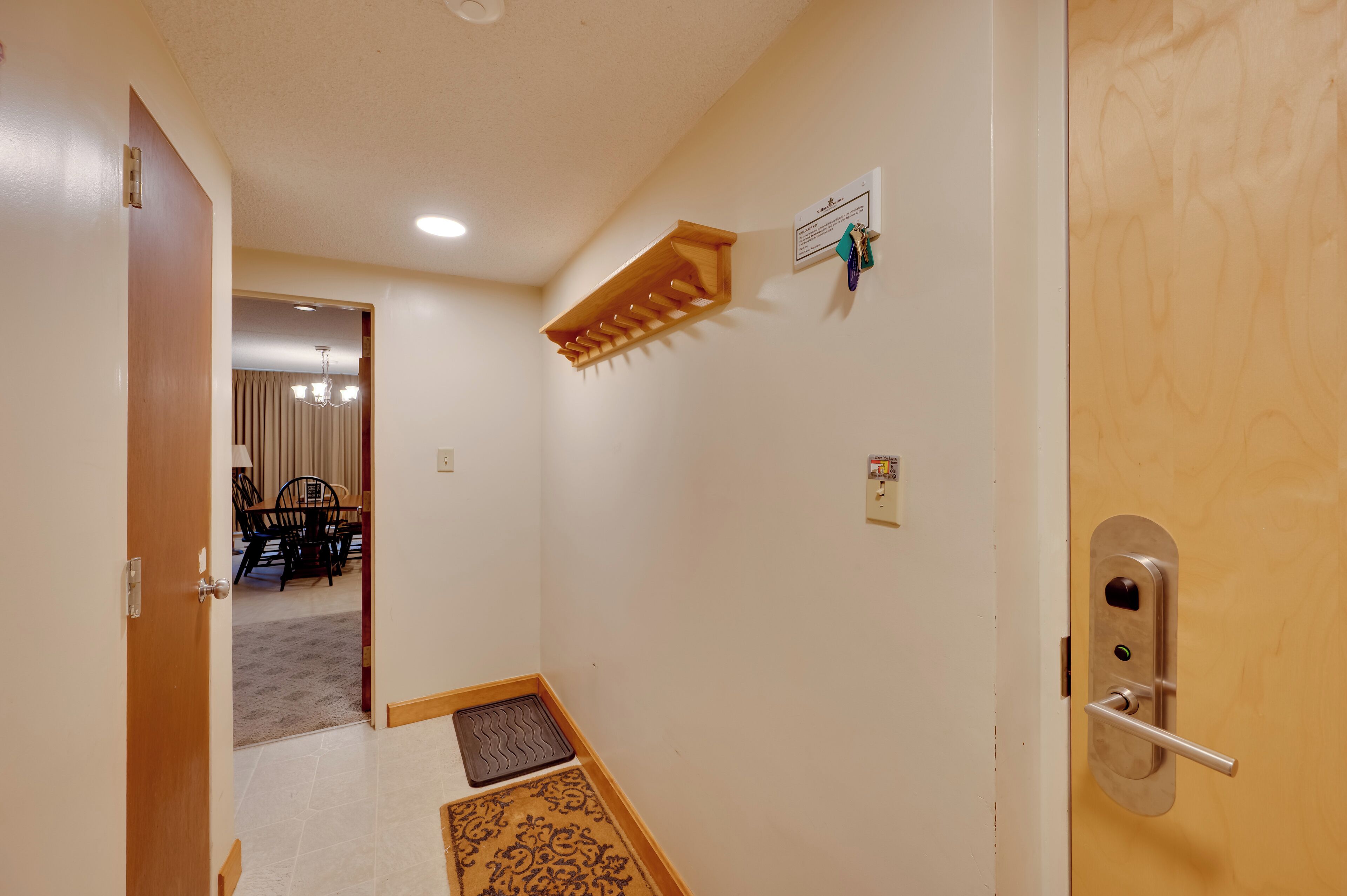 Un pasillo con una puerta de madera, un equipo de aire acondicionado instalado en la pared, un timbre y un felpudo en la entrada.