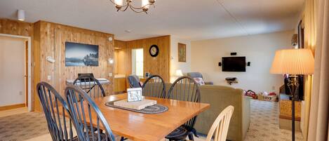 A dining area with a wooden table and chairs, a chandelier, and a television in the background.