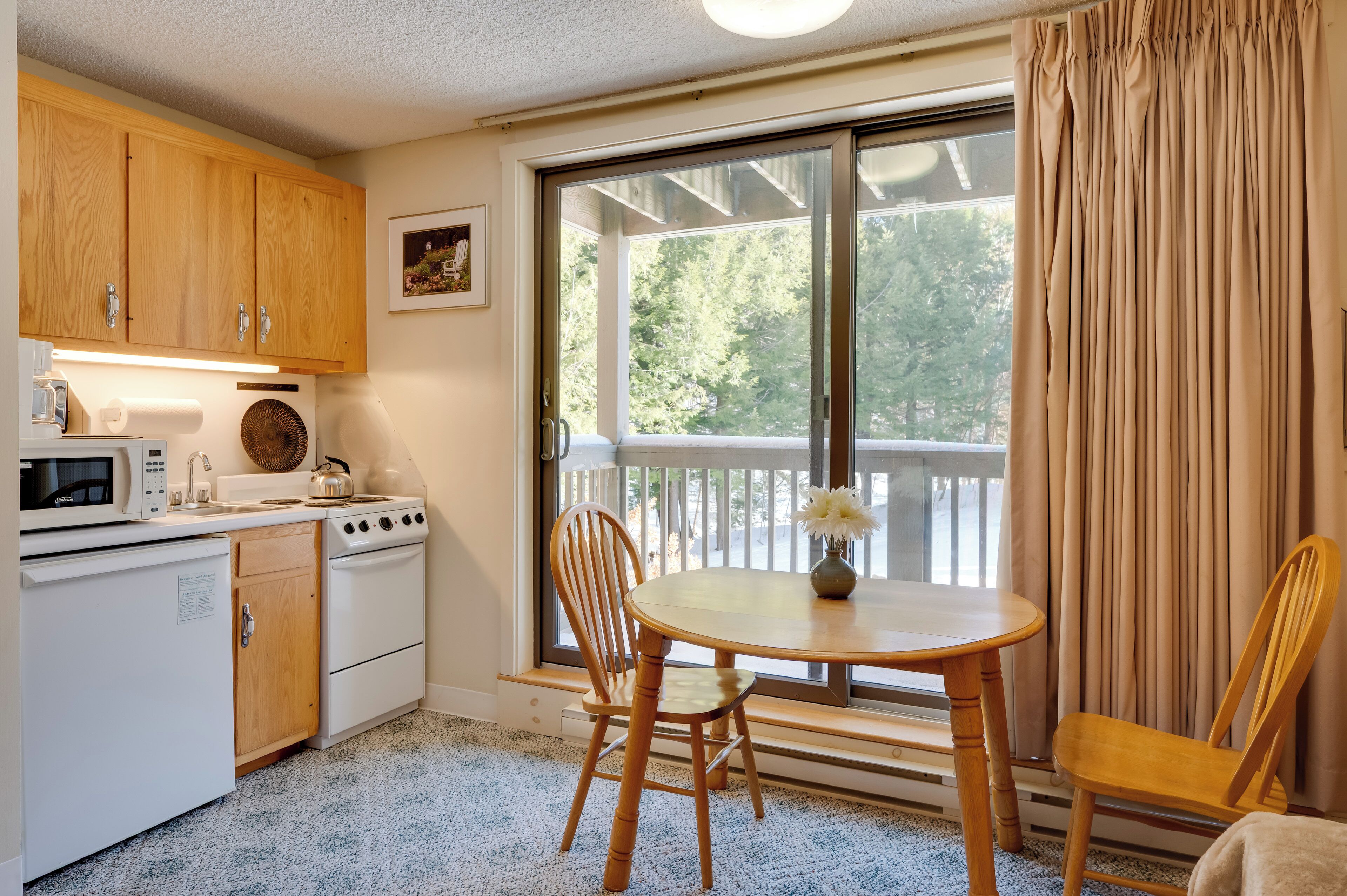 A compact kitchen with wooden cabinets, a stove, and a microwave. There is a dining table with two chairs and a window with curtains.