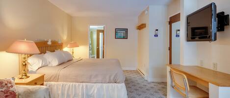 A bedroom with a bed, bedside lamps, a desk with a chair, a TV, and a door leading to another room.