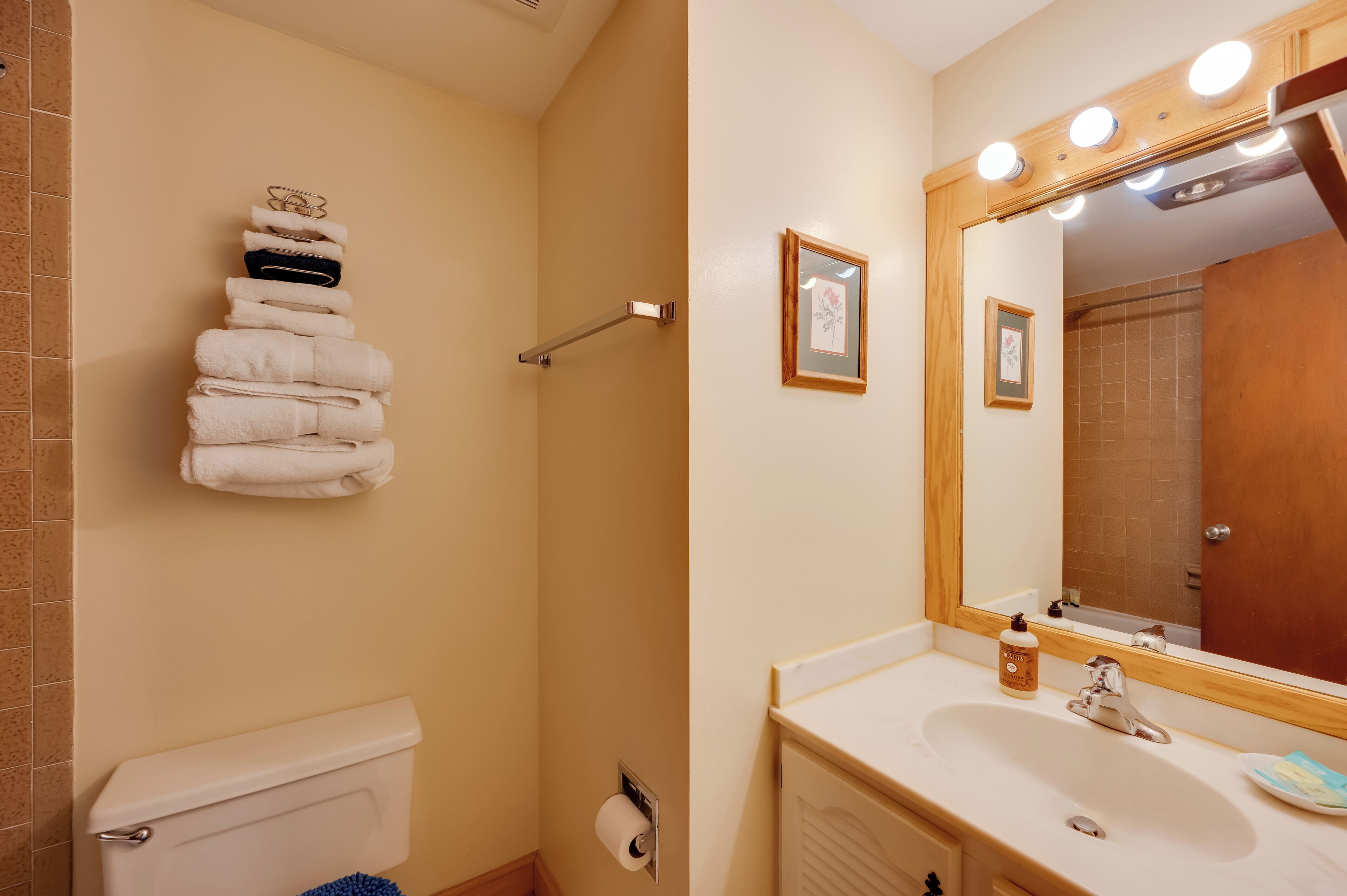 A bathroom with a toilet, a stack of white towels, a shower, a sink with a mirror, and a framed picture on the wall.