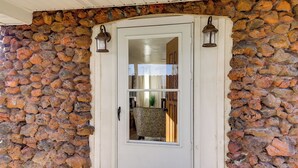 A stone wall entrance with a white door, a small window, and two wall-mounted lanterns. - Clear Lake Living! Pet-friendly Gem w/ Fenced Yard (Lakeport)