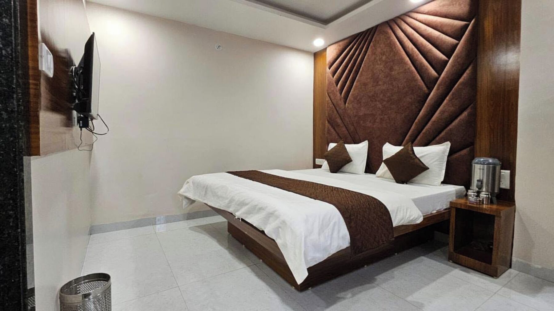 Habitación de hotel con una cama grande, cabecera de madera, mesita de noche con lámpara y televisor montado en la pared.