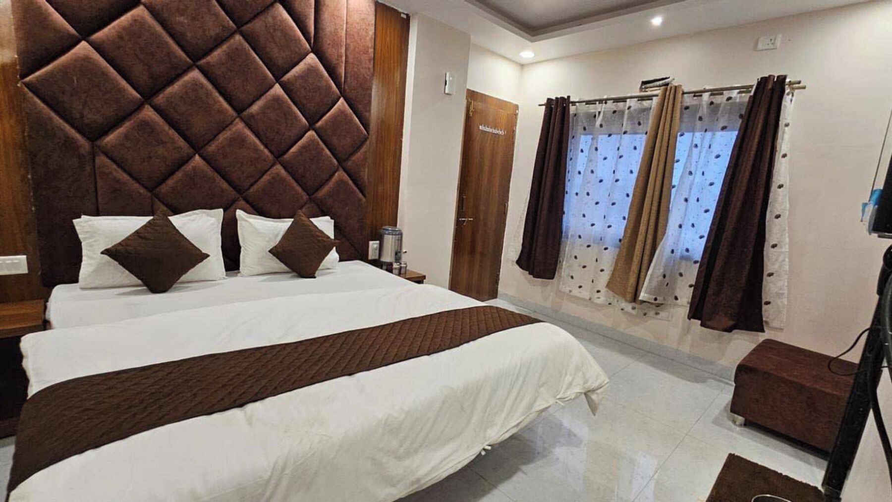 A hotel room with a large bed, a wooden headboard, a bedside table, a small bedside table, a brown ottoman, and a window with curtains.