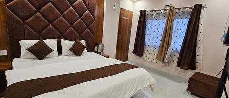 A hotel room with a large bed, a wooden headboard, a small bedside table, a brown ottoman, and a window with curtains.