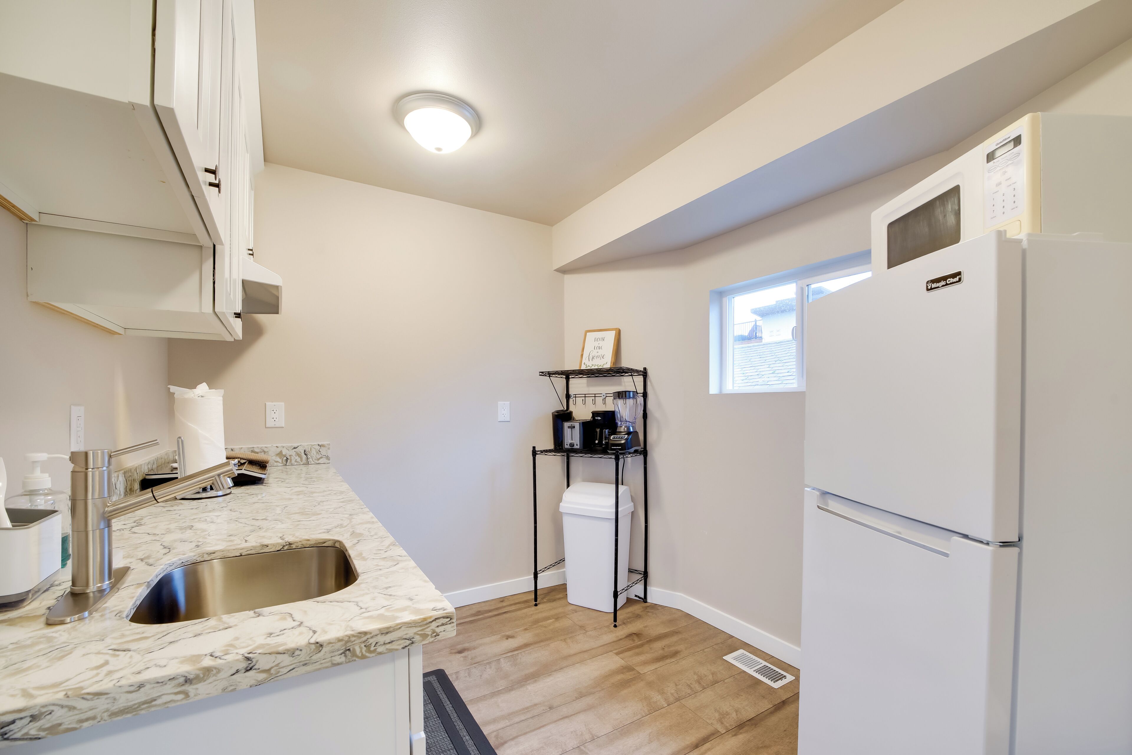 A modern kitchen with a marble countertop, stainless steel sink, white cabinets, a black shelving unit, and a white refrigerator.