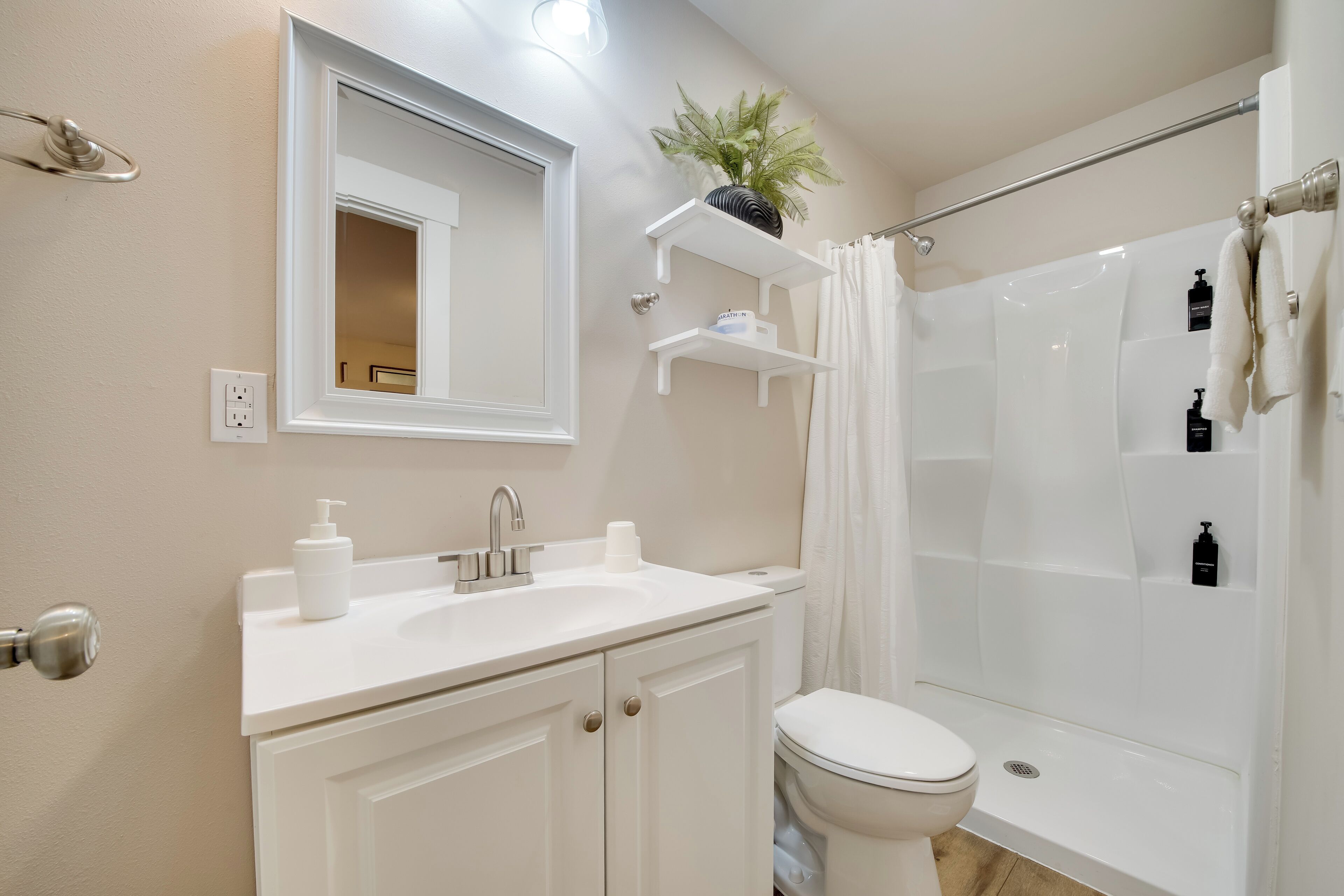 A modern bathroom with a white sink, toilet, and shower. There are white cabinets, a mirror, and a plant on a shelf.