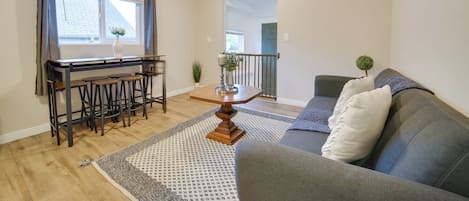 A living room with a grey sofa, wooden dining table, and bar stools.