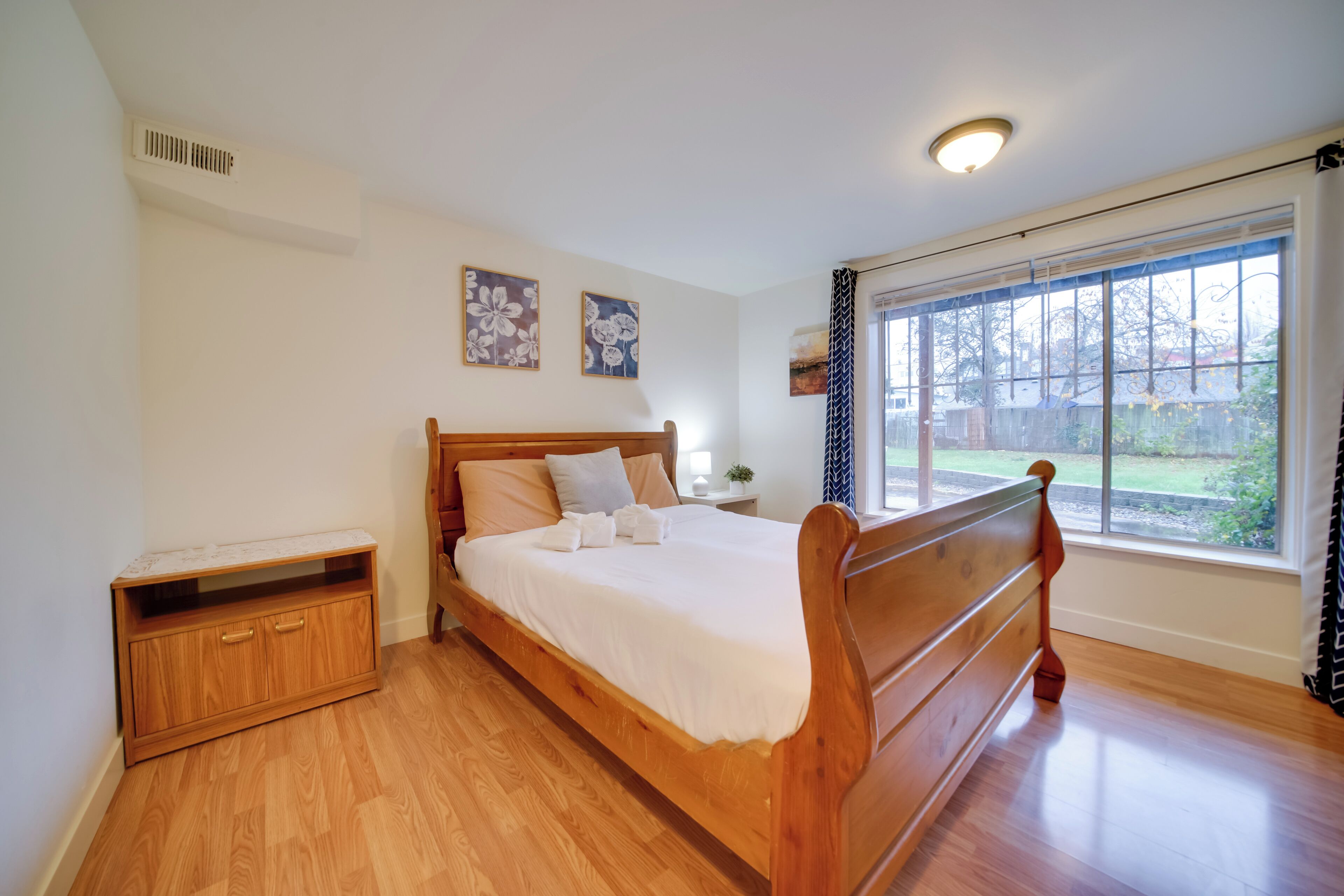 Ein Schlafzimmer mit einem hölzernen Bett, einem Nachttisch, einer kleinen Kommode und einem Fenster mit Blick ins Freie.