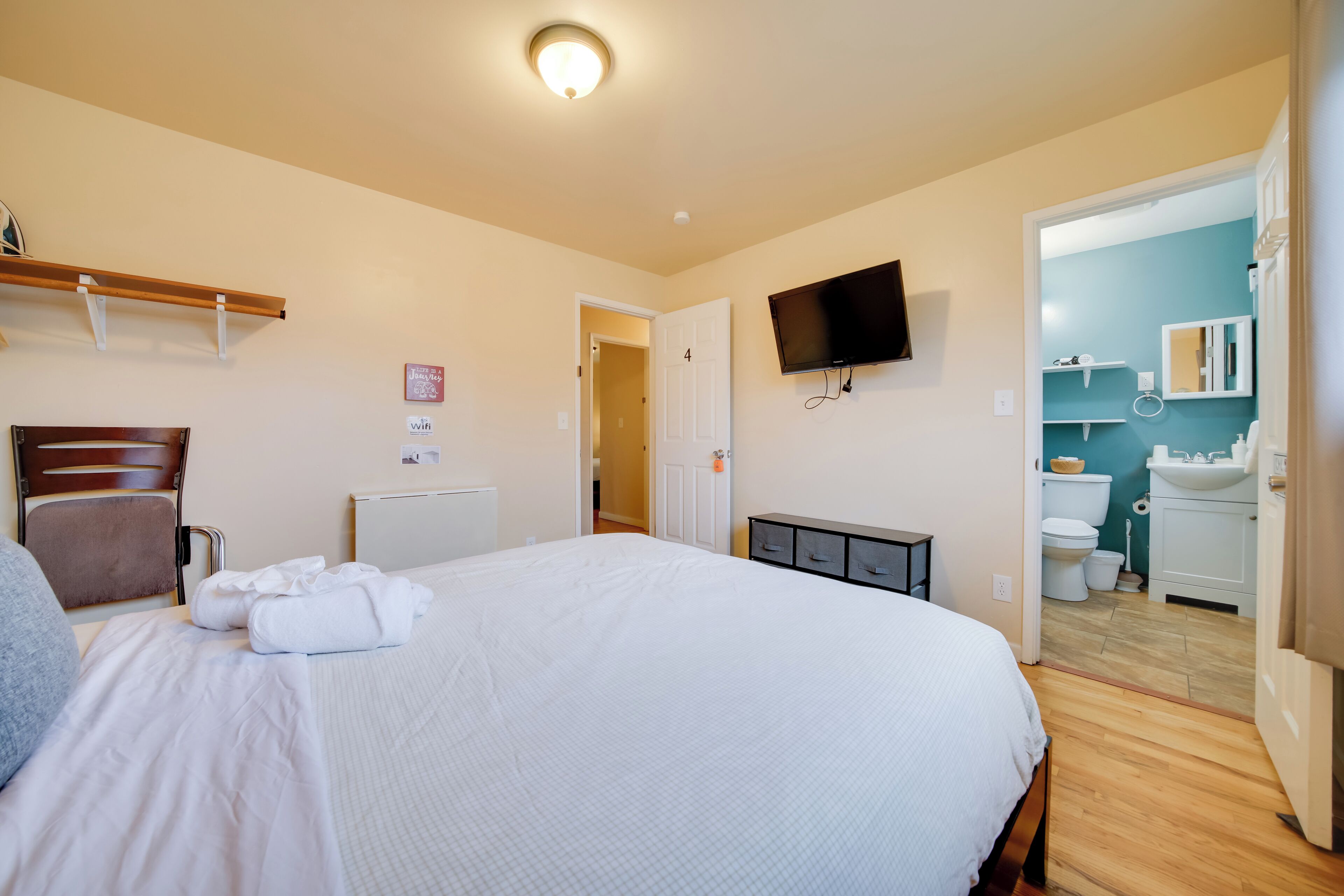 A hotel room with a large bed, a TV mounted on the wall, a wooden headboard, a bedside table, and a door leading to the bathroom.