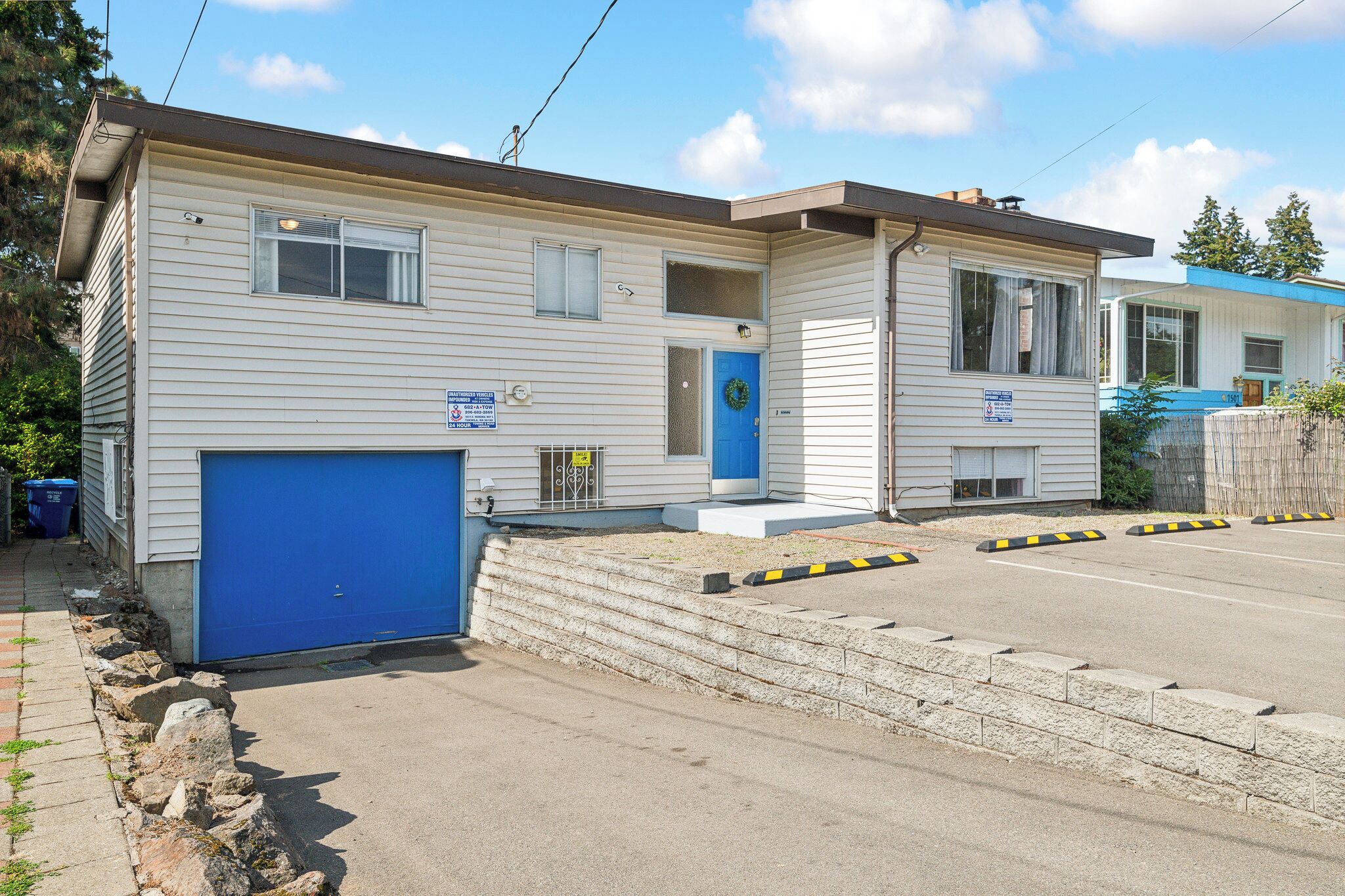 Une maison mobile à deux étages, avec une porte de garage bleue, un petit porche et un jardin devant la maison, bordé d’un muret en pierre.