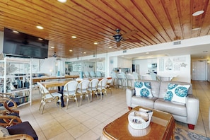A spacious living area with a wooden ceiling, a flat-screen TV, a dining area with white chairs, and a sofa with patterned cushions. - Beachfront 3BR w/ Pools, Hot Tubs & Sauna (South Padre Island)