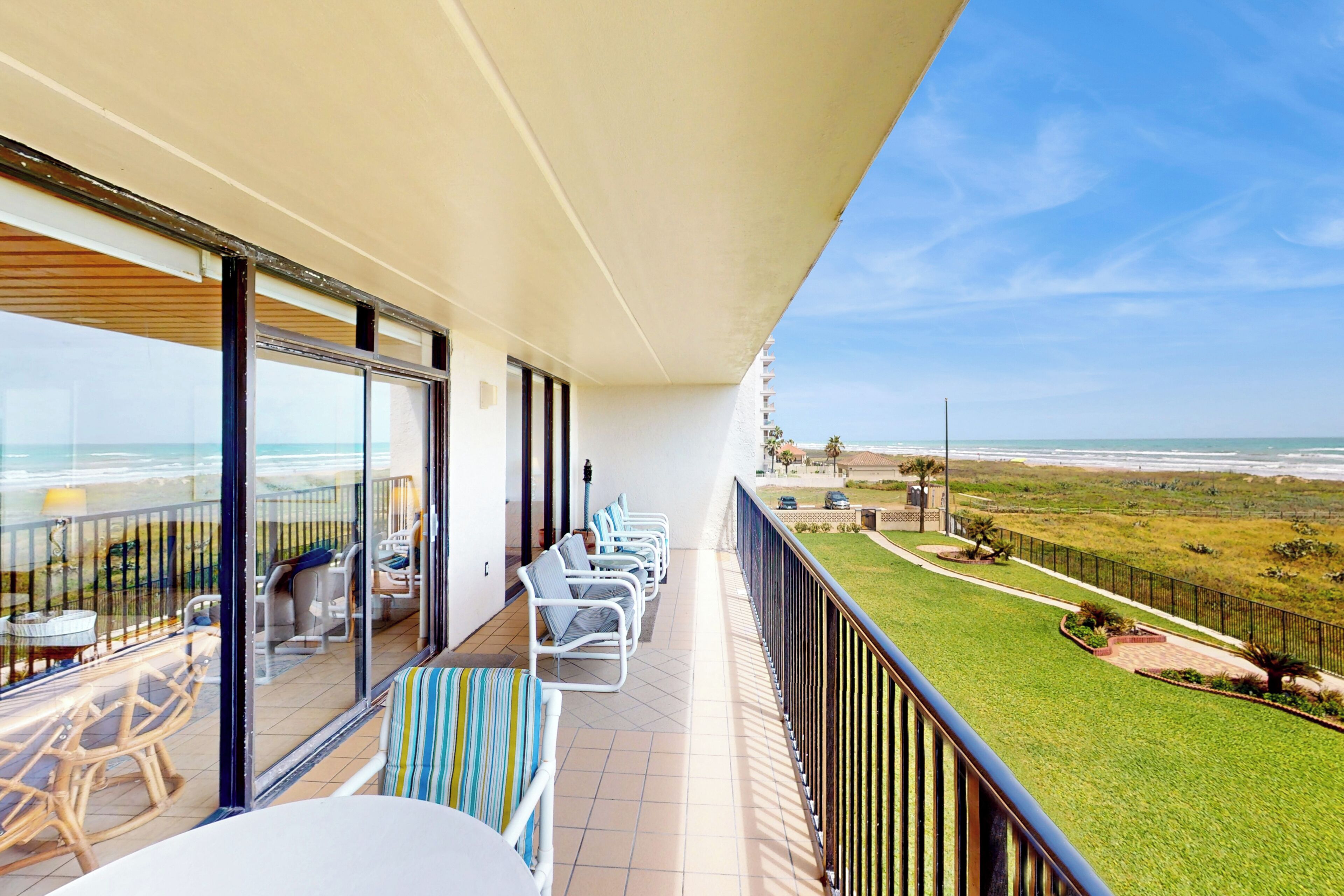 Ein Balkon mit Blick auf den Strand, ausgestattet mit Stühlen und Tischen, und eine Rasenfläche mit einer Umzäunung.
