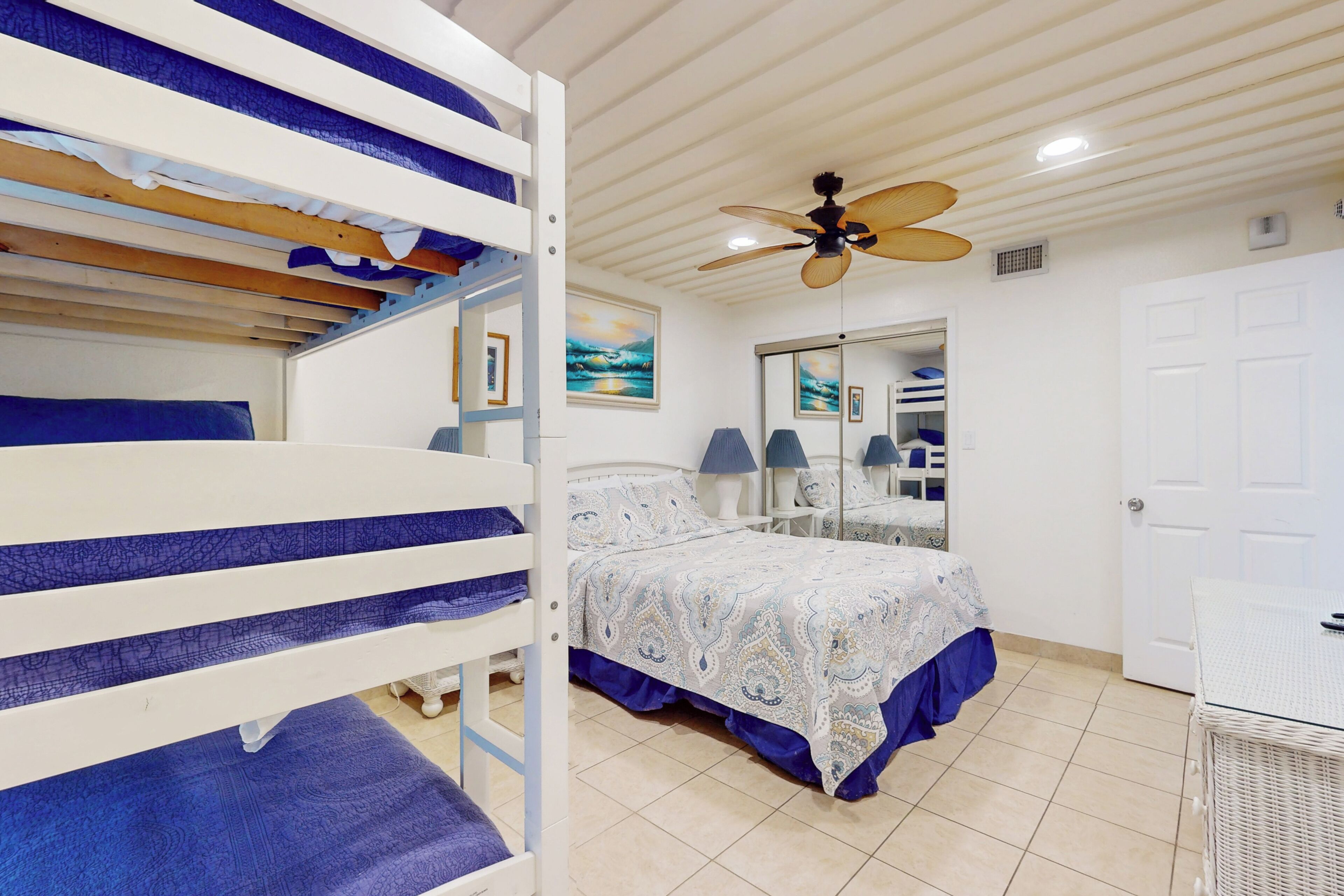 A bedroom with bunk beds, a ceiling fan, a bed with a patterned bedspread, a mirror, and a door.