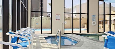 Zona de piscina interior con paredes de vidrio transparente, una piscina y sillones de sala reclinables en azul y blanco.