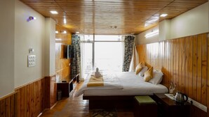 A hotel room with wooden paneling, a large bed with pillows, a bedside table with a kettle, and a view through a large window. - Hotel Pride (Kalimpong)