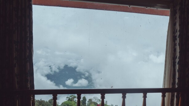 A room with a wooden floor, a green table with a tray of cups and a flower vase, and a view of the sky and greenery outside. - Hotel Pride (Kalimpong)