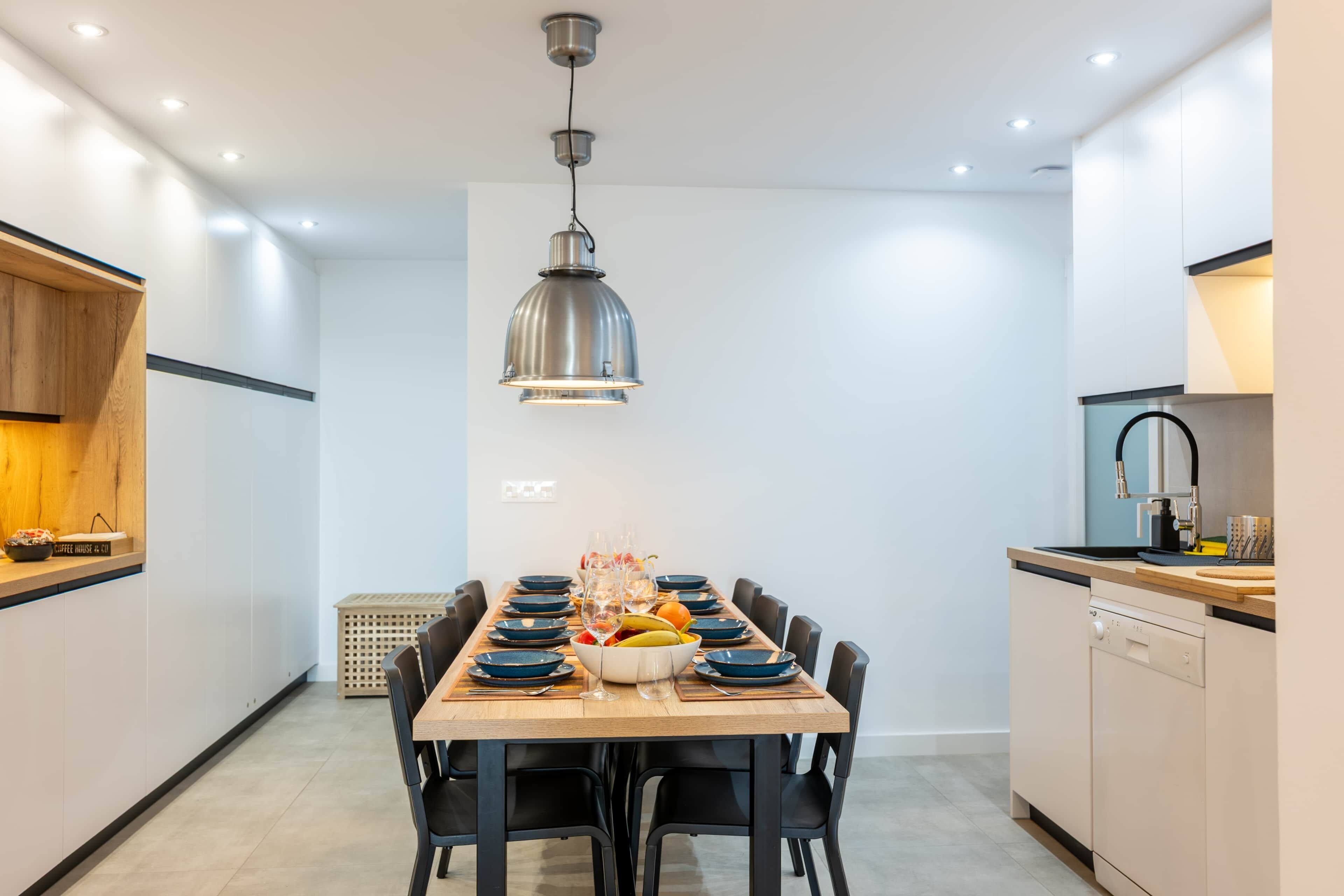 A modern kitchen with a dining table set for six, equipped with black chairs, a central island, and a pendant light.