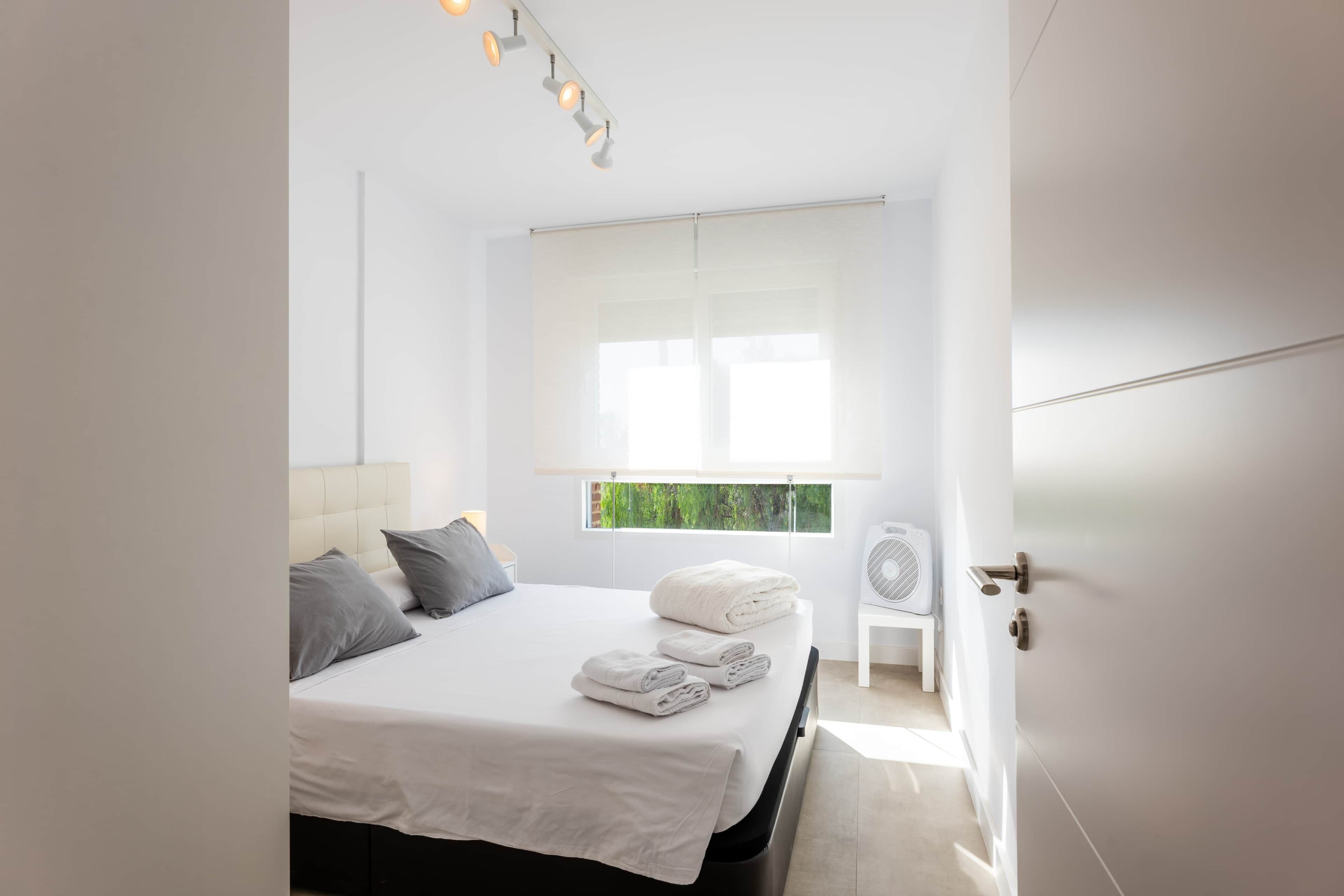 Une chambre à coucher moderne avec un lit, des oreillers, une fenêtre avec des stores, un ventilateur de chevet et une porte.