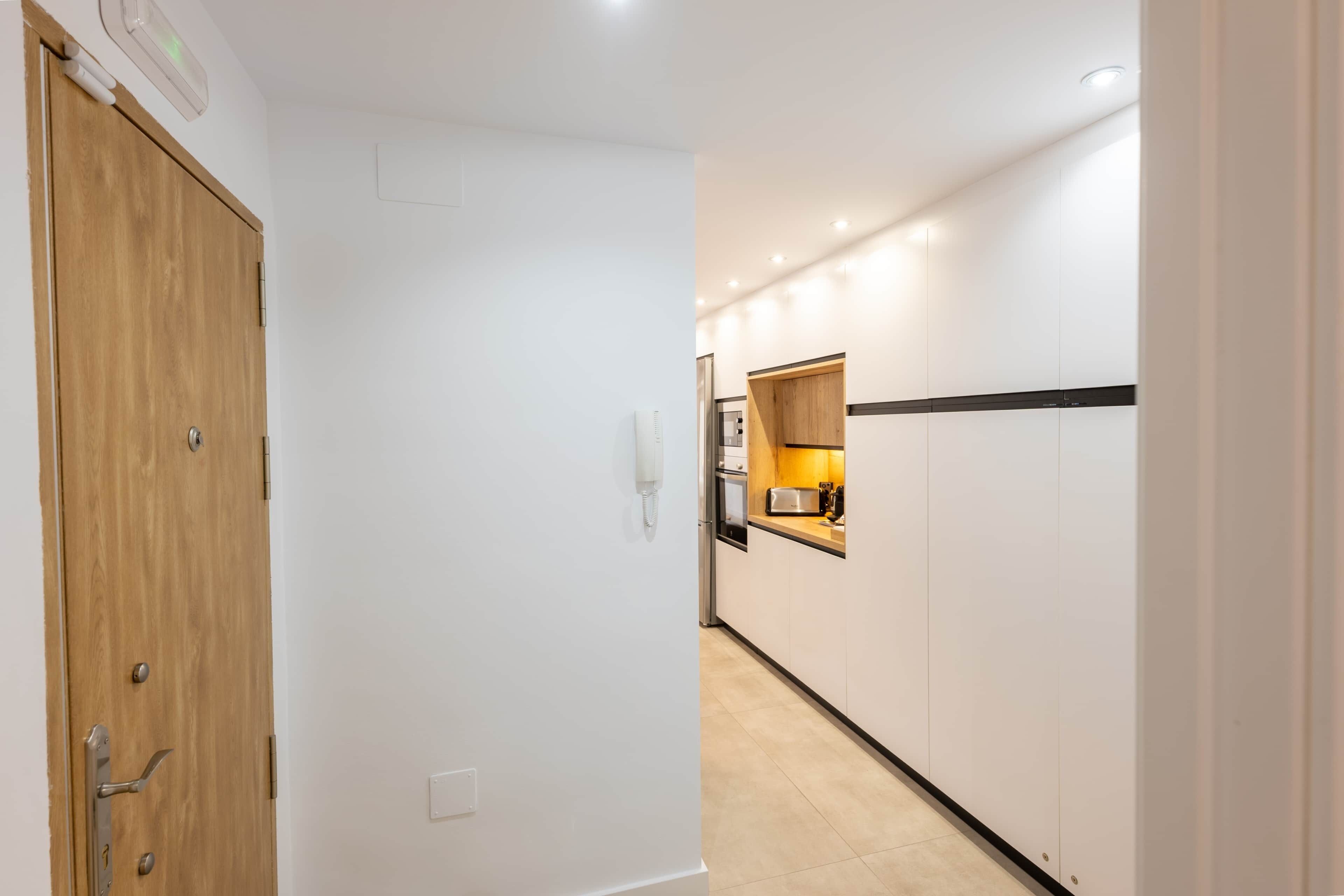 A hallway with a wooden door, a white wall, and a kitchen area with cabinets and a countertop.