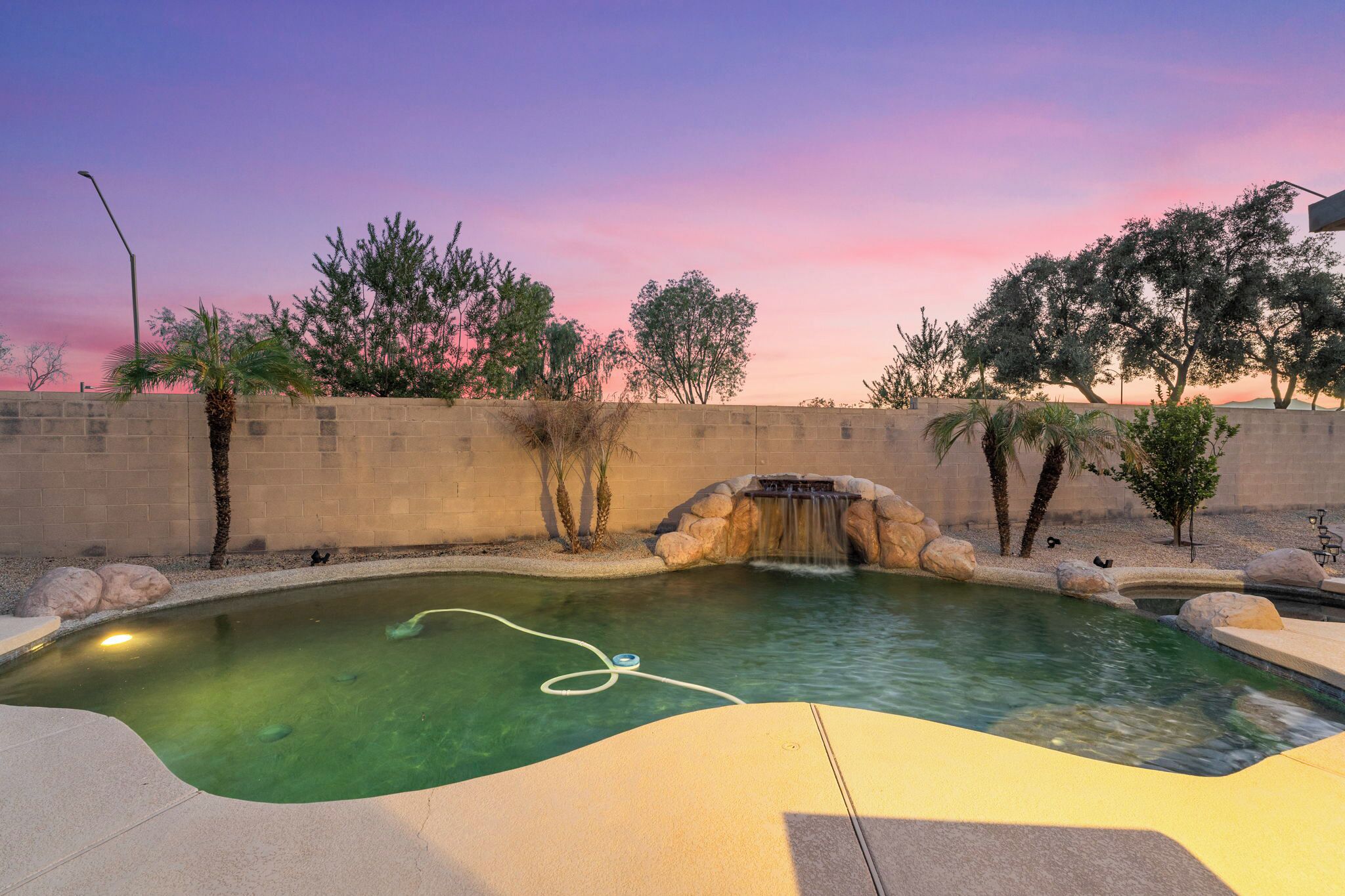 A swimming pool with a waterfall feature, surrounded by a low stone wall and various trees.