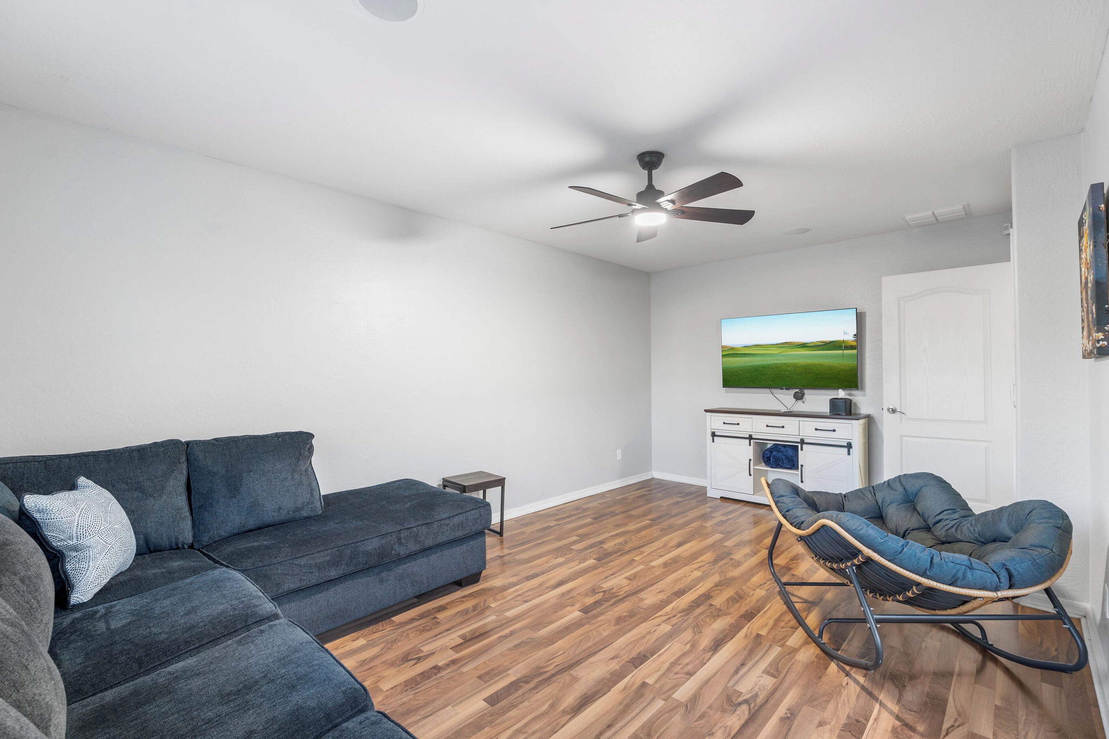 A modern living room with a dark grey sectional sofa, a rocking chair, a flat-screen TV, and a ceiling fan.